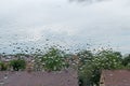 Raindrops on window glass make pattern on background of blue cloudy sky and blurred brown roofs and greenery of trees below