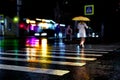Rainy city street reflections. Unrecognizible woman with yellow umbrella cross street by crosswalk. City life in night in rain