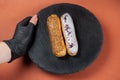 Raspberry and caramel eclair on round black rock stand held by hand in black glove. Eclairs with glaze in cafe. View