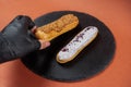 Raspberry eclair and caramel eclair held by hand in black glove on round black rock stand. Eclairs with glaze in cafe