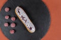 Raspberry eclair on round black rock stand on brown background. Eclair with white glaze and raspberries in cafe. View