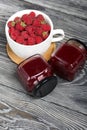 Raspberry jam in glass jars. Nearby are raspberries in a large white mug. On wooden boards with a beautiful texture