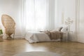 Rattan peacock chair next to window in white bedroom interior with blanket on bed