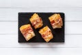 Ravioli wrapped in fried bacon slices, canape on black stone plate, white wooden table.