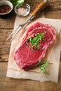 Raw beef steak. Fresh beef rib eye steak with fork rosemary, salt and pepper on piece of parchment paper on old wooden background. Royalty Free Stock Photo