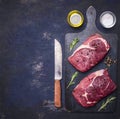 Raw beef steak with rosemary,butter and meat fork on dark rustic cutting board, top view with space for tex