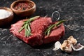 Raw beef steak with rosemary near garlic cloves and small bowls with salt and peppercorns on black marble surface.