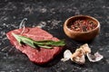 Raw beef steak with rosemary twig near small bowl with peppercorns mix and garlic on black marble surface. Royalty Free Stock Photo