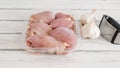 Raw chicken thighs in a plastic container, and fresh garlic and garlic press close-up on a white wooden board with copy space Royalty Free Stock Photo