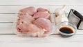 Raw chicken thighs in a plastic container, soy sauce, and fresh garlic and garlic press close-up on a white wooden board Royalty Free Stock Photo