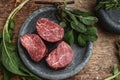 3 raw fillet mignon steaks in grey stone plate on rustic wood table with fresh green herbs top view.