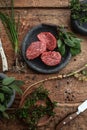 3 raw fillet mignon steaks in grey stone plate on rustic wood table with steak knifes and fresh green herbs top view.