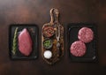 Raw fresh beef sirloin fillet steak and mince burgers sealed in vacuum tray on dark kitchen background with spices and fork