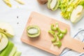 Raw green ingredients for smoothie, whole and cut apples, chopped celery stalk on a wooden board on white cloth background Royalty Free Stock Photo