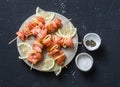 Raw ingredients for cooking salmon skewers on the grill. Raw salmon skewers, lemon, pepper, sea salt on a cutting board on dark ba