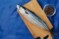 Raw mackerel fish whole, on a cutting board, bowl with a mixture of peppers and spices and a blue satin tablecloth. Top view with