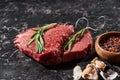 Raw meat steaks with rosemary twigs near small bowl with peppercorns and garlic on black marble surface. Royalty Free Stock Photo
