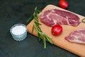 Raw pork steak, rosemary, tomatoes and salt on wooden cutting board, dark background, top view and side view Royalty Free Stock Photo