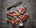 Raw t-bone steaks, butcher`s knife, seasoning, and grilled lemon halves on gray concrete textured background, top view