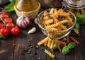 Raw tricolor fusilli pasta in glass bowl with oil and garlic, basil plant and tomatoes with pepper and linen towel on wooden Royalty Free Stock Photo