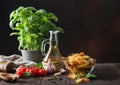 Raw tricolor fusilli pasta in glass bowl with oil and garlic, basil plant and tomatoes on wooden background Royalty Free Stock Photo