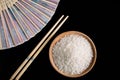 Raw white rice, hand fan and chopsticks on black background. Long uncooked rice in a wooden plate.