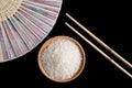 Raw white rice, hand fan and chopsticks on black background. Long uncooked rice in a wooden plate.