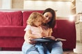 Reading big book. Young mother with her little daughter in casual clothes together indoors at home Royalty Free Stock Photo