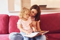 Reading big book. Young mother with her little daughter in casual clothes together indoors at home Royalty Free Stock Photo