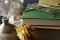 READING GLASSES AND CASE ON TWO BOOKS IN AN ARRANGEMENT WITH A DRY ROSE, FLUFFY FEATHER, GLASS LAMP AND CERAMIC VASE