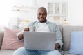 Reading morning news. Happy african american man using laptop and drinking hot coffee, resting on sofa Royalty Free Stock Photo