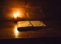 Reading scene in ancient times: an old book leaning on ruined wooden table lighted by a candle on a wooden background Royalty Free Stock Photo