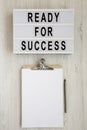 `Ready for success` words on a lightbox, clipboard with blank sheet of paper on a white wooden background, top view. Overhead, Royalty Free Stock Photo