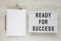 `Ready for success` words on a lightbox, clipboard with blank sheet of paper on a white wooden background, top view. Overhead, Royalty Free Stock Photo