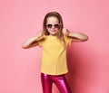 Blonde kid girl in yellow t-shirt, shiny pink leggings and sunglasses holds hands up with fists clenched