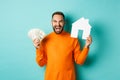 Real estate concept and mortgage concept. Ecstatic caucasian man holding money dollars and house maket, shouting for joy
