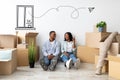 Real estate concept. Young african american couple sitting near wall among cardboard boxes, woman pointing at free space Royalty Free Stock Photo