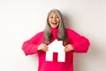 Real estate. Excited asian senior woman looking happy and lucky, winning apartment, showing paper house model and Royalty Free Stock Photo