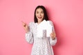 Real estate. Excited asian woman showing paper house model, pointing and looking left at copy space, standing over pink Royalty Free Stock Photo