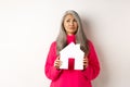 Real estate. Sad asian senior woman dreaming about own property, holding paper house model and grimacing upset, looking Royalty Free Stock Photo