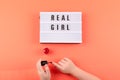 Real girl text on lightbox and girl hands paints nails with red nail polish on coral background. Beauty, makeup flat lay