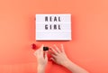 Real girl text on lightbox and girl hands paints nails with red nail polish on coral background. Beauty, makeup flat lay