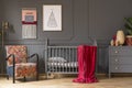 Real photo of a baby crib with a red blanket, an armchair, lamp