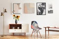 Real photo of bright eclectic living room interior with many posters, colorful chair, wooden cupboard with flowers and coffee tabl