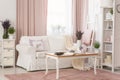 Real photo of bright provencal sitting room interior with white sofa, wooden coffee table on dirty pink carpet, rack with decor an