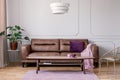 Real photo of brown leather sofa with violet cushion and pastel pink blanket standing in light grey sitting room interior with Mon Royalty Free Stock Photo