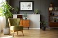 Real photo of dark living room interior with green armchair, vintage cupboards, fresh plants and poster on wall with wainscoting Royalty Free Stock Photo