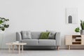 Real photo of grey lounge with green cushion, wooden coffee table, simple poster on the wall and cupboard with books in bright sit