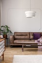 Real photo of modern lamp hanging above leather couch with purple pillow in bright living room interior with coffee table, molding