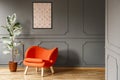 Real photo of open space dark grey living room interior with poster hanging on wall with wainscoting, orange armchair and fresh p